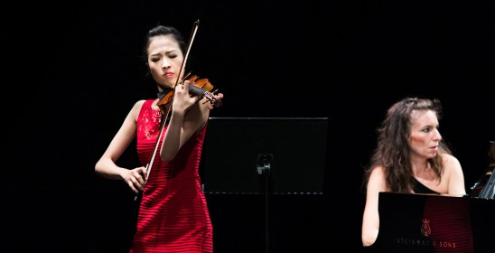 Kobieta w czierwonej sukni grająca na skrzypcach, obok kobieta grająca na fortepianie